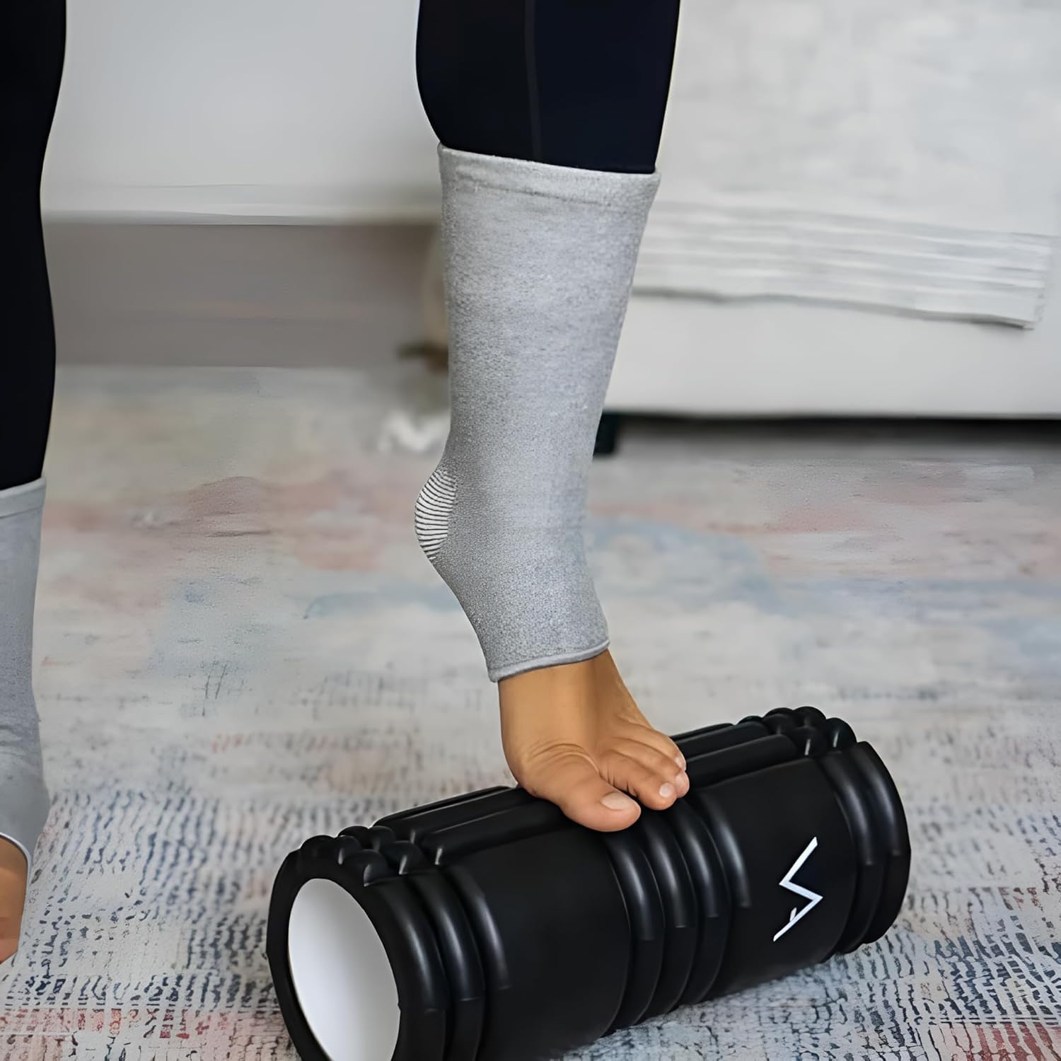 Person using black foam roller, wearing grey ankle brace, on textured rug indoors